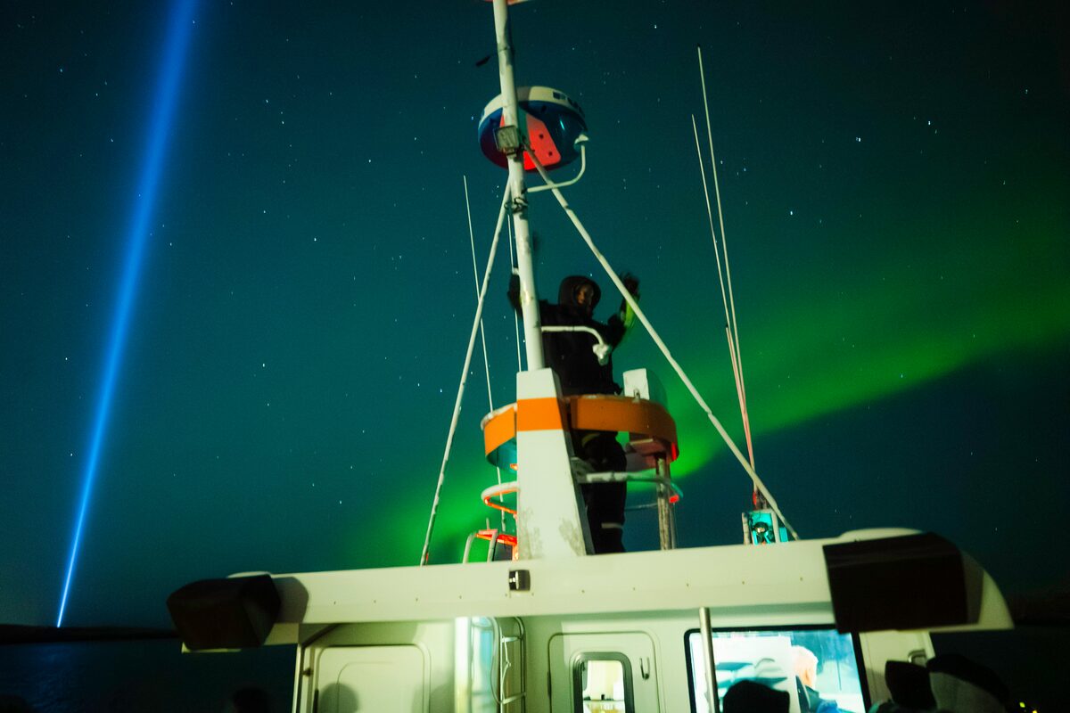 Person standing on a ship mast under an aurora.