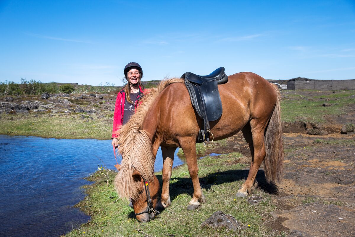Smiling woman next to a saddled horse drinking from a pond.