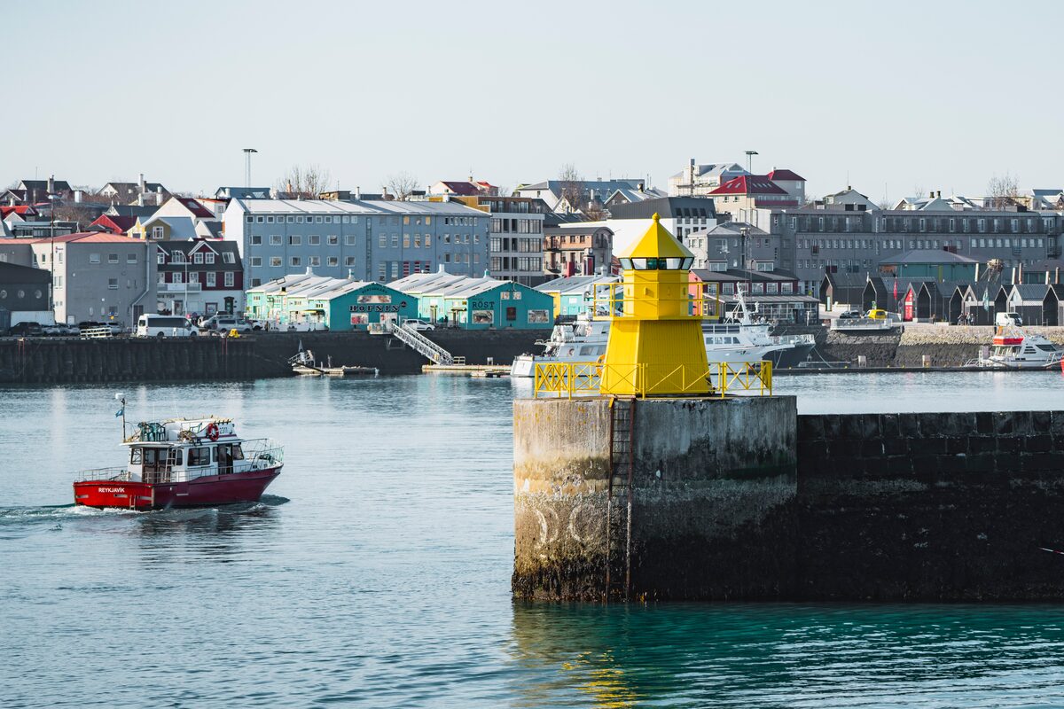 A small red boat near a yellow lighthouse in a harbor.